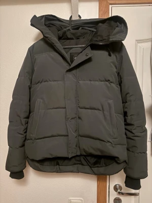 Mörkgrå pufferjacka från Canada Goose black label - Mörkgrå pufferjacka från Canada Goose med huva och snyggt broderat logomärke på ärmen. Jackan har dragkedja och knappstängning, två sidofickor och ribbade muddar vid ärmslut. Perfekt för kalla dagar med sin dunfyllda insida och stilrena look.