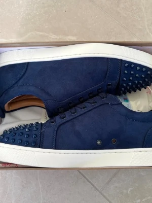 Mörkblå sneakers med nitar - Säljer ett par mörkblå sneakers från Christian Louboutin med coola nitar på tån och sidorna. Skorna har snörning, vit sula och röd undersida. Tillverkade i mocka med detaljer i skinn på insidan. Perfekt för dig som gillar att sticka ut med din stil.