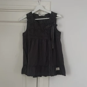 Svart blus med spets från Odd Molly - Supersöt mörkgrå blus från Odd Molly med broderad spets upptill och en rosett vid axeln. Blusen är ärmlös och har volangkant nedtill.