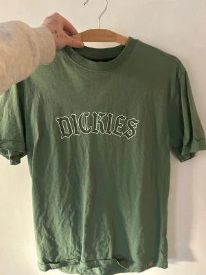 Grön Dickies t-shirt med tryck - Säljer en grön t-shirt från Dickies med stort vitt tryck på bröstet. Klassisk rund hals och korta ärmar, perfekt för chill dagar. Materialet är mjuk bomull som känns skönt mot huden. Enkel och stilren design som funkar till jeans eller shorts.
