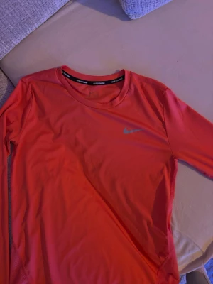 Nike Dri-Fit löpartröja orange - Snygg orange långärmad löpartröja från Nike med Dri-Fit-teknologi. Tröjan har rund hals och ett diskret Nike Swoosh på bröstet. Perfekt för dig som gillar att träna och vill ha något som andas och sitter skönt. Svart band med 'NIKE RUNNING' i nacken.