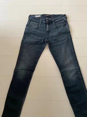 Replay Anbass mörkblå jeans - Snygga Replay Anbass jeans i mörkblå tvätt med klassisk femficksdesign och diskreta slitningar. Modellen har smal passform och raka ben, med detaljerade sömmar på bakfickorna och logotyp på myntfickan. Perfekt för dig som gillar stilrena och moderna jeans. 30W 32L