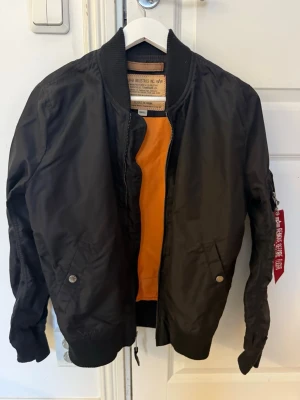 Svart bomberjacka Alpha Industries - Säljer en svart bomberjacka från Alpha Industries med klassisk orange insida och coolt 'Remove Before Flight'-tag på ärmen. Jackan har dragkedja, ribbade muddar och flera fickor. Perfekt för dig som vill ha en streetig och tidlös look.