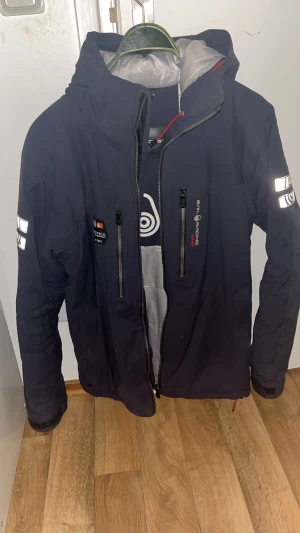 Mörkblå vindjacka Sail Racing M - Snygg mörkblå vindjacka från Sail Racing med huva, reflekterande detaljer på ärmarna och flera dragkedjor framtill. Jackan har coola loggor och patchar på bröstet och är tillverkad i slitstark polyester. Ett spänne/armjustering vid handleden saknas. Jackan fungerar som vanligt i övrigt. Säljs i befintligt skick. Privatförsäljning – ingen retur.