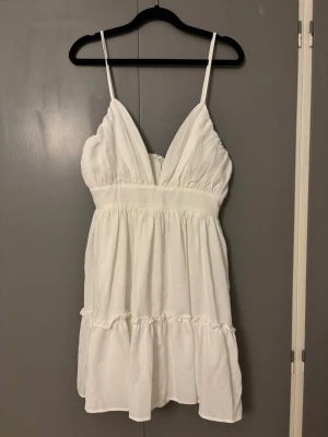 Vit kort klänning från PrettyLittleThing - Supersöt vit klänning med smala axelband och v-ringad urringning från PrettyLittleThing. Modellen är kort med volangdetaljer och har dragkedja i ryggen. Perfekt för sommarens alla tillfällen, lätt och luftig i polyester.