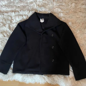 Svart kappa - Klassisk svart kappa med bred krage och dubbelknäppning framtill. Jackan är tillverkad i ull och har en rak, boxig passform med långa ärmar. Perfekt för dig som gillar en stilren och tidlös look. Från Gina Young storlek 170 men sitter som en xs-s