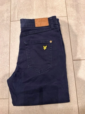Mörkblå byxor från Lyle & Scott Junior - Snygga mörkblå byxor från Lyle & Scott Junior med klassisk passform. Byxorna har fem fickor, varav en med den ikoniska gula fågeln broderad. Tillverkade i mjuk bomull med detaljer i brunt läder vid linningen och metallknappar. Perfekta för dig som gillar stilrena och bekväma plagg.