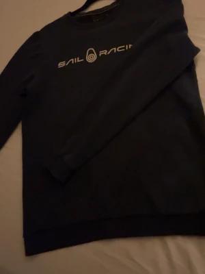Blå Sail Racing sweatshirt - Blå sweatshirt från Sail Racing med vit logotyp tryckt på bröstet. Tröjan har rund hals, långa ärmar och ribbade muddar vid ärmslut och nederkant. Perfekt för dig som gillar stilrena och sportiga plagg.