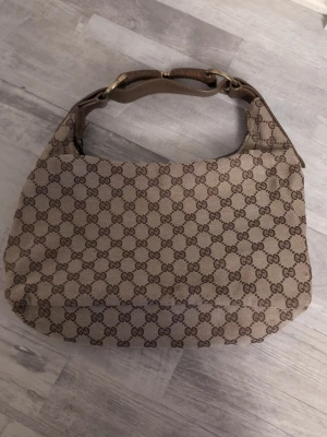 Gucci beige axelväska med GG-mönster - Säljer en beige axelväska från Gucci med det klassiska GG-mönstret i brunt. Väskan har detaljer i brunt skinn, guldfärgade metalldelar och dragkedja upptill. Perfekt storlek för det viktigaste och en ikonisk accessoar för dig som gillar designer.