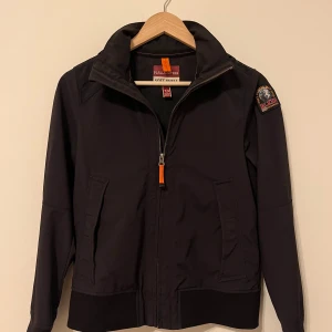 Svart soft shell jacka Parajumpers - Svart soft shell jacka från Parajumpers med huva, dragkedja och två fickor framtill. Snygg patch på ärmen och orange detalj vid dragkedjan. Jackan har en sportig look och är perfekt för dig som gillar stilrena och funktionella plagg.