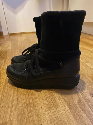 Svarta moon boots med snörning - Säljer ett par svarta moon boots med snörning och rund tå. Skorna har en mjuk pälsfodrad insida och är tillverkade i mocka och skinn. Perfekta för kalla dagar med sin höga och fluffiga design. Snygga och bekväma att dra på sig när det är vinter.