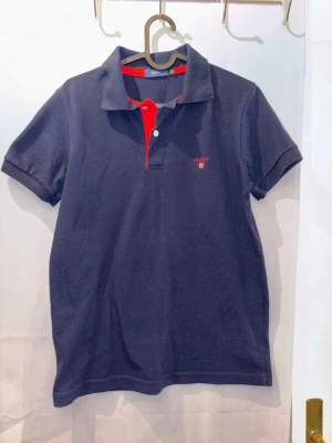 Mörkblå pikétröja från Gant - Klassisk mörkblå pikétröja från Gant med röd knappslå och broderad logga på bröstet. Tröjan har korta ärmar och krage, perfekt för en stilren look. Materialet känns mjukt och bekvämt.