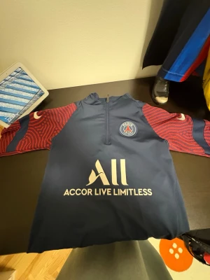 PSG träningströja blå/röd Nike - Snygg långärmad träningströja från PSG i marinblått med röda mönstrade ärmar. Tröjan har halv dragkedja, klubbmärke på bröstet och Accor Live Limitless-tryck framtill. Perfekt för fotbollsträning eller sportiga dagar.