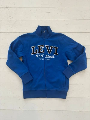 Vintage Blå Zip Sweatshirt  Levi Finland - Vintage Blå ziptröja från Levi med hög krage och tryck framtill där det står 'LEVI 67,8° North Finland'. Tröjan har långa ärmar, ribbade muddar och dragkedja hela vägen. Perfekt för en avslappnad och sportig stil.