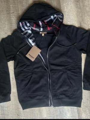 Svart hoodie från Burberry - Svart hoodie med dragkedja från Burberry. Klassisk rutig insida på huvan i svart, vitt och rött. Två stora fickor framtill och ribbade muddar. Perfekt för en stilren och trendig look.