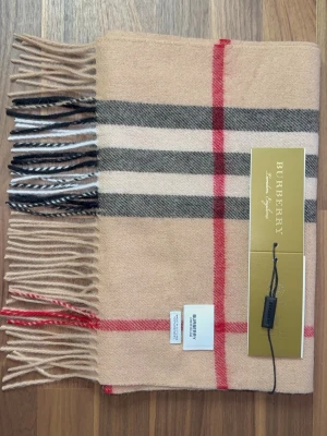 Burberry Halsduk - Klassisk beige halsduk från Burberry i 100% kashmir med det ikoniska rutmönstret i svart, vitt och rött. Halsduken har fransar i ändarna och känns både lyxig och tidlös. Perfekt accessoar för att lyfta vilken outfit som helst. Halsduken är oanvänd(ny). Skriv vid minsta lilla fundering!