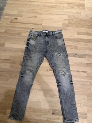 Grå slitna skinny jeans - Säljer ett par grå skinny jeans med slitningar och tvättad look. Jeansen har klassisk femficksmodell, bälteshällor och dragkedjegylf. Perfekta för dig som gillar en edgy och trendig stil.
