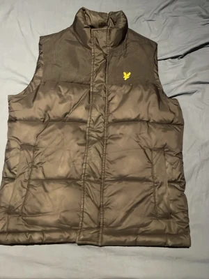 Svart dunväst från Lyle & Scott - Snygg svart dunväst från Lyle & Scott med gul logga på bröstet. Västen har hög krage, quiltad design och dragkedja framtill. Perfekt för lager på lager och håller dig varm under kyliga dagar. Tillverkad i glansigt material med klassisk passform.