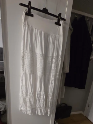 Vit långkjol med spetsdetaljer - Säljer en vit långkjol med bred midjeresår och flera sektioner med fina spetsdetaljer. Kjolen har en luftig och flowy känsla, perfekt för varmare dagar. Passar till många olika stilar och är superenkel att matcha.