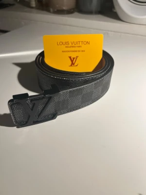 Louis Vuitton svart bälte med LV-spänne - Snyggt svart bälte från Louis Vuitton med klassiskt LV-monogram och ett stort LV-spänne i svart metall. Bältet är gjort i präglat läder och har en stilren, modern look som passar till jeans eller kostymbyxor.
