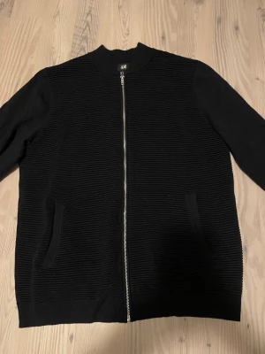 Marinblå ribbad cardigan jacka H&M - Svart cardigan jacka från H&M med ribbad struktur på fram- och baksida. Jackan har hel dragkedja framtill, två fickor och långärmade släta ärmar. Perfekt för en stilren och enkel look.