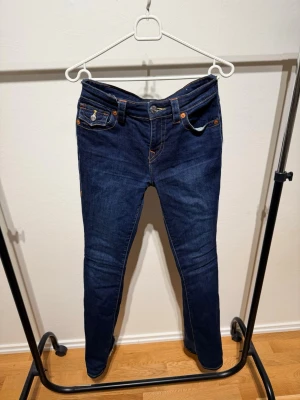 Mörkblå jeans från True Religion - Snygga mörkblå jeans från True Religion med klassiska kontrastsömmar och unika bakfickor med lock och knapp. Jeansen har låg midja och utsvängande modell är Becca, Perfekta för dig som vill ha en cool och tidlös look. Ska även nämnas att byxorna var lite för långa så det är lite slitet längst ner men resten är  bra skick.