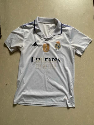 Real Madrid tröja vit  - Säljer en vit Real Madrid matchtröja från Adidas med klubbmärke, Adidas-logga och FIFA-märke på bröstet. Tröjan har korta ärmar, krage och blå detaljer på axlarna. Tillverkad i lätt och ventilerande material, perfekt för fotbollsträning eller match.