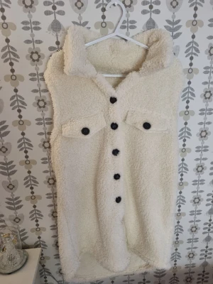Vit teddyväst med stora knappar - Mysig vit teddyväst i fuskpäls med stora svarta knappar framtill och två bröstfickor med lock. Västen har en hög krage och är ärmlös, perfekt för lager på lager och en trendig look.