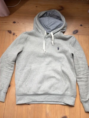 Grå hoodie från Polo Ralph Lauren - Snygg ljusgrå hoodie från Polo Ralph Lauren med klassisk huva och vita dragsnören. Tröjan har en broderad logga på bröstet och en stor magficka. Tillverkad i mjukt bomullsmaterial för extra komfort. Perfekt för en chill och avslappnad stil.