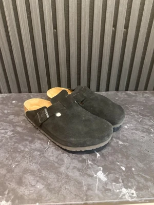 Svarta Birkenstock tofflor i mocka - Säljer ett par svarta Birkenstock tofflor i mjuk mocka med klassisk rem och spänne. De har en bekväm innersula i kork och en slitstark yttersula. Perfekta för dig som vill ha både stil och komfort i vardagen.