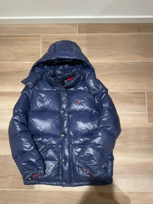 Blå Ralph Lauren Puffer Jacka  - Mörkblå dunjacka från Polo Ralph Lauren med huva och röd logga på bröstet. Jackan har quiltad design, tryckknappar och dragkedja framtill samt elastiska muddar. Perfekt för kalla dagar och riktigt snygg till vintern.