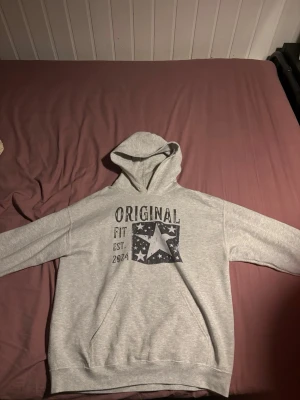 Grå hoodie med tryck och huva - Snygg grå hoodie med svart tryck framtill där det står 'ORIGINAL FIT EST. 2024' och en stjärna omgiven av små stjärnor. Hoodien har en klassisk huva och känguruficka framtill. Perfekt för en avslappnad stil.