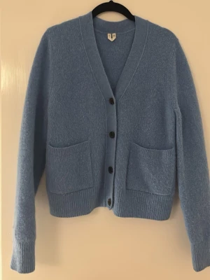 Arket - Ljusblå kofta med fickor och knappar - Mysig ljusblå kofta med v-ringning och fyra svarta knappar framtill. Koftan har två stora fickor på framsidan och ribbade muddar vid ärmslut och nederkant. Perfekt för lager på lager och enkel att matcha med olika outfits.