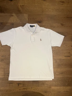 Vit pikétröja från Polo Ralph Lauren - Klassisk vit pikétröja från Polo Ralph Lauren med korta ärmar och krage. Tröjan har knappar vid halsen och den ikoniska flerfärgade broderade loggan på bröstet. Tillverkad i mjuk bomull för en bekväm känsla och clean look.