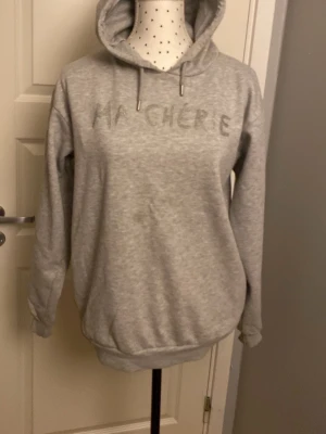 Grå hoodie med text Ma Chérie - Säljer en ljusgrå hoodie med huva och dragsko. På bröstet står det 'MA CHÉRIE' i glittriga stenar. Hoodien har ribbade muddar vid ärmar och nederkant. Perfekt för en chill och avslappnad stil. En liten fläck på magen. Den är även lite nopprig