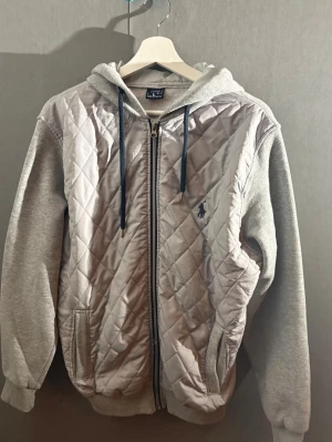 Grå quiltad hoodie från Ralph Lauren - Snygg grå hoodie från Ralph Lauren med quiltad framsida och dragkedja. Har huva med mörka snören och broderad logga på bröstet. Ärmar och baksida i mjuk sweatshirtmaterial, perfekt för en avslappnad men stilren look.