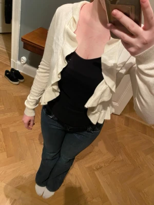 Krämvit volangkofta - Söt krämvit kofta med volangkant framtill. Kortare modell med långa ärmar och mjukt, stickat material. Perfekt att slänga över ett linne eller t-shirt för en extra touch.