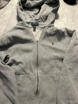 Grå hoodie från Polo Ralph Lauren - Snygg grå hoodie från Polo Ralph Lauren med dragkedja framtill och klassisk blå logga på bröstet. Mjuk fleece på insidan och stor huva. Perfekt för chill dagar eller när du vill ha en enkel men stilren look. Två fickor framtill och ribbade muddar. Den är i storlek xs passar runt 165. Pris kan diskuteras vid snabb affär!