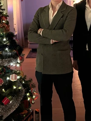 John hendric kavaj  - Stilren mörkgrön kavaj med dubbelknäppning och sex bruna knappar framtill. Kavajen har tre utanpåliggande fickor och klassisk krage. Materialet ser ut att vara mjukt och något stretchigt, perfekt för en dressad men avslappnad look.