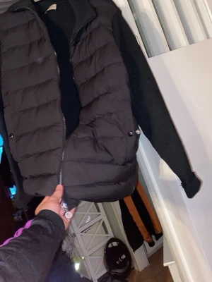 Svart cardigan jacka från Moncler - Säljer en svart cardigan jacka från Moncler med quiltad front och stickade ärmar. Jackan har dragkedja framtill, två fickor med knappstängning och Moncler-logga på ärmen. Perfekt för dig som vill ha en stilren och bekväm jacka.