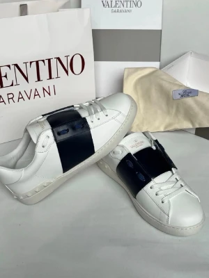 Valentino Garavani Open sneakers vit/blå - HELT NYA!! Snygga Valentino Garavani Open sneakers i vitt skinn med bred mörkblå rand över mitten. Klassisk låg modell med rund tå, vita snören och diskret logga på plösen. Perfekta för dig som vill ha en clean men ändå unik look. Först till kvarn!