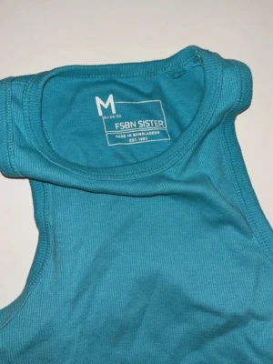 Turkos ribbad linnetopp FSBN Sister - Snygg turkos ribbad linnetopp från FSBN Sister. Modellen är croppad och ärmlös med rund halsringning. Perfekt för varma dagar eller som layering-piece. Materialet är mjukt och stretchigt för en bekväm passform.