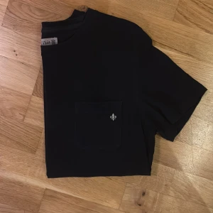 Mörkblå t-shirt från Morris med ficka - Mörkblå t-shirt från Morris i storlek XS. T-shirten har en klassisk passform med korta ärmar och en rund hals. På bröstet finns en ficka med en broderad vit lilja som detalj. Materialet är mjuk bomull som känns skön mot huden.