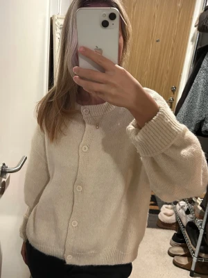 Beige stickad kofta - Mysig krämvit stickad kofta med rund halsringning. Koftan har ribbade muddar och knäpps med fem matchande knappar framtill. Perfekt för lager på lager och enkel att styla till olika outfits.