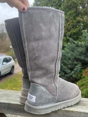 Gråa UGG Australia mockastövlar - Klassiska gråa UGG-stövlar i mjuk mocka med fårskinnsfoder. Höga skaft och rund tå ger en mysig och avslappnad look. Perfekta för kalla dagar och riktigt sköna att ha på sig. UGG-logga bak på hälen.