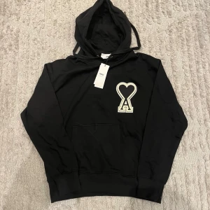 Svart hoodie från Ami Paris med logga - Svart hoodie från Ami Paris med stor vit broderad hjärtlogga på bröstet. Klassisk känguruficka framtill, ribbade muddar och justerbar huva med snören. Perfekt för en avslappnad och trendig look.