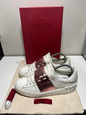 Valentino Garavani sneakers vit/rosa - Snygga vita sneakers från Valentino Garavani med en metallicrosa rem över mitten. Skorna har klassisk snörning, rund tå och detaljerade nitar på hälen. Tillverkade i skinn för en lyxig känsla och look. Bra skick i storlek 38, dustbag och snören ingår