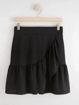 Svart omlottkjol med volang - Snygg svart kortkjol med omlottdesign och volangkant. Kjolen har en bred midja och en dekorativ detalj framtill som ger en trendig look. Perfekt att matcha med en enkel topp eller stickad tröja.