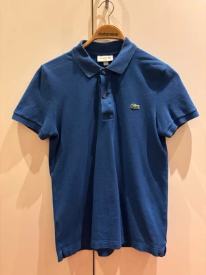 Blå pikétröja från Lacoste - Klassisk blå pikétröja från Lacoste med korta ärmar och krage. Tröjan har knappar framtill och den ikoniska krokodilloggan på bröstet. Perfekt för en stilren och sportig look.