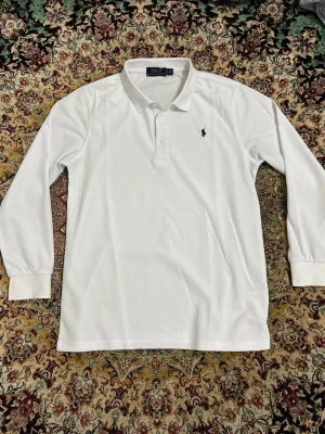Vit långärmad piké från Polo Ralph Lauren - Klassisk vit långärmad pikétröja från Polo Ralph Lauren med broderad logga på bröstet. Tröjan har krage, knappar framtill och är tillverkad i mjukt bomullsmaterial. Perfekt för en clean och stilren look.